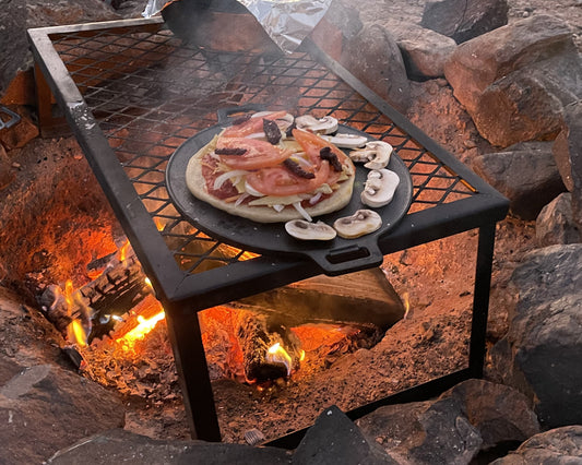 A pizza on a skillet is being cooked on a steel grate over a wood campfire.