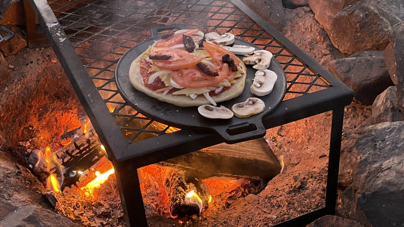 A pizza on a skillet is being cooked on a steel grate over a wood campfire.